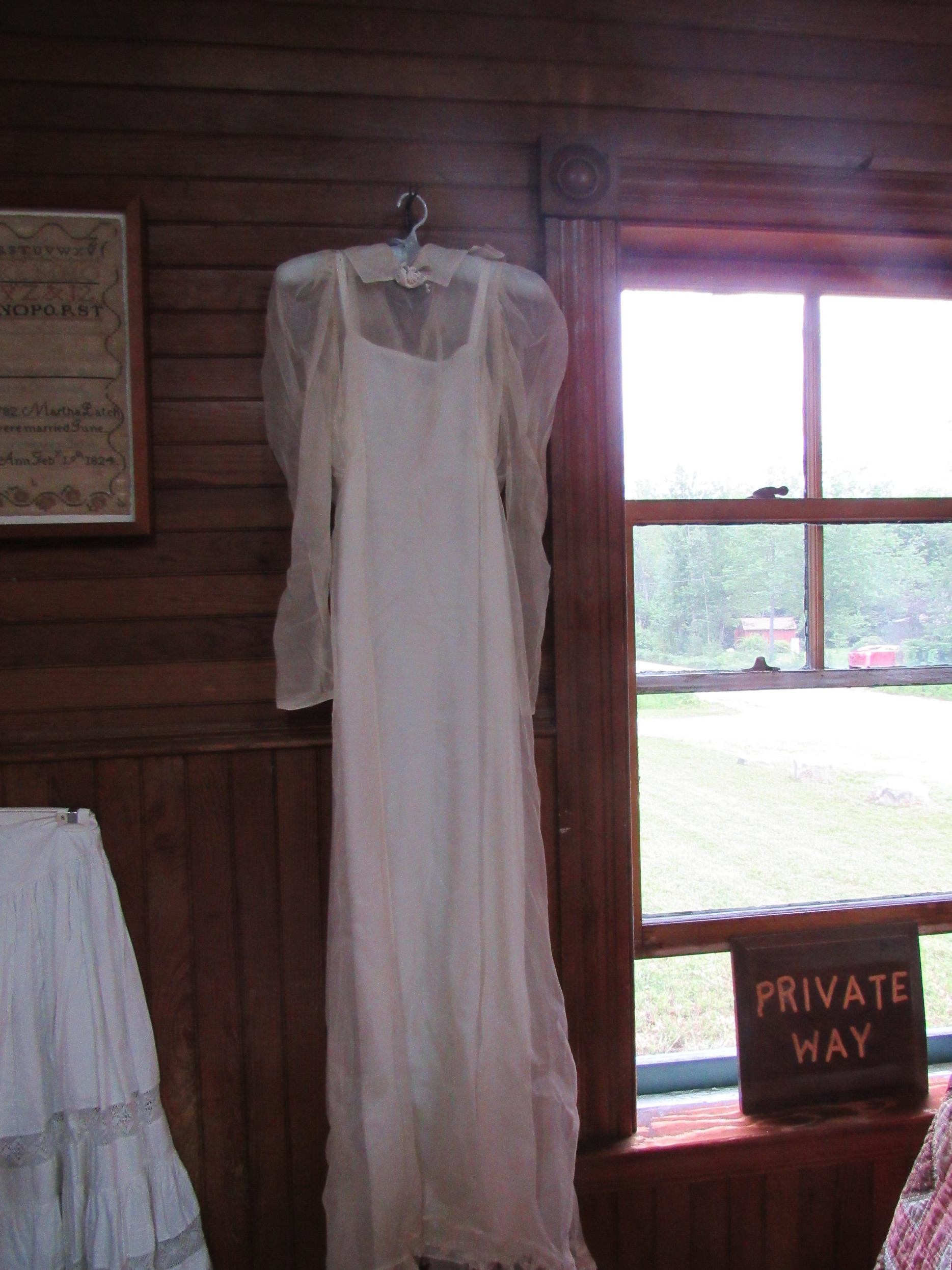 Long white dress beside a framed needlework sampler.