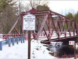 Red metal truss bridge over the Crooked River, with snow on the banks and a Ryefield Bridge sign.